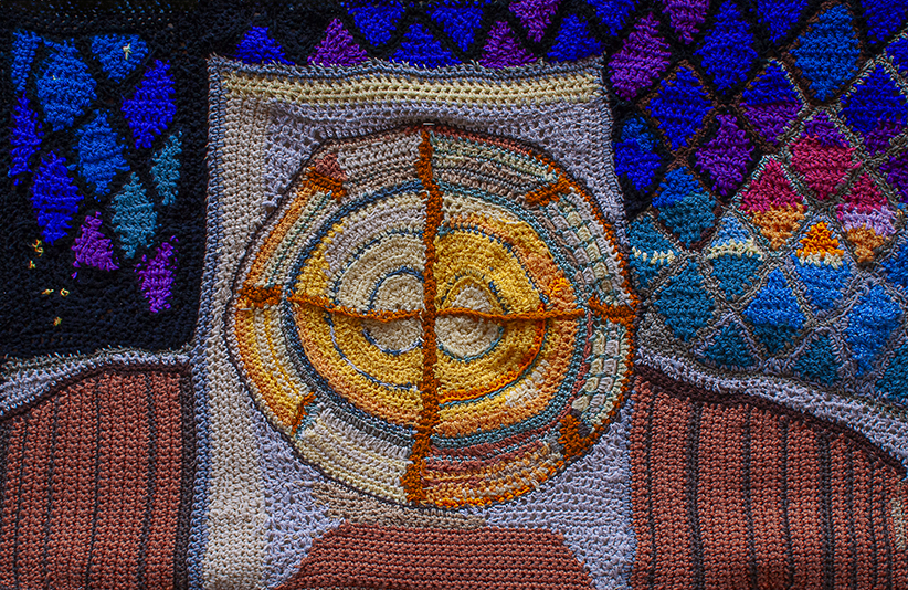 A detailed crocheted representation of a lighthouse lens surrounded by colorful geometric patterns, showcasing a blend of textures and colors.