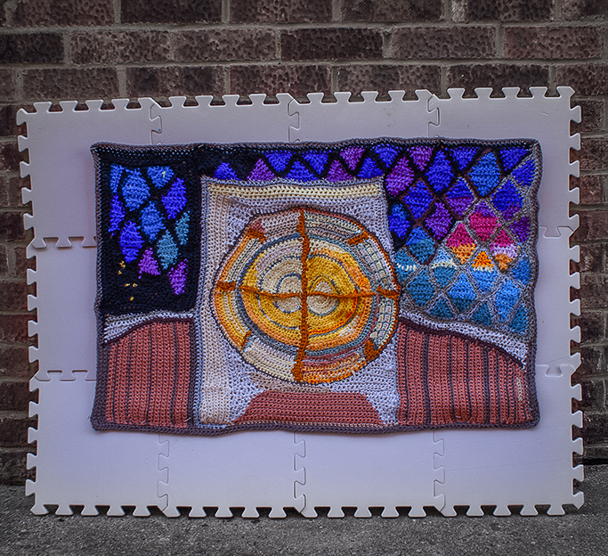 A crocheted artwork depicting the lens of a lighthouse with vibrant geometric patterns, set against a textured background.