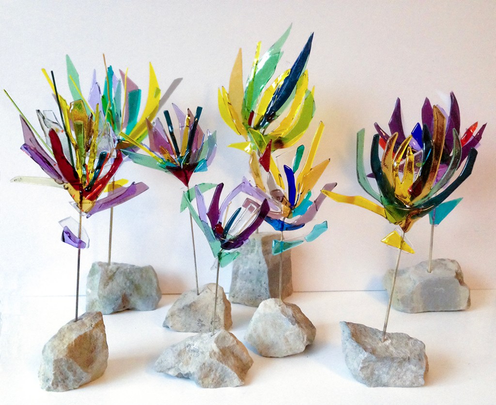 A collection of colorful glass flowers mounted on thin metal rods, each emerging from small rocks, arranged artfully against a plain background.