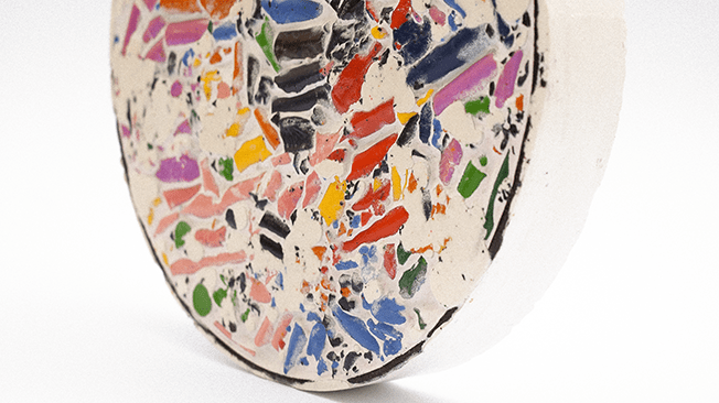 A close-up view of a colorful, mosaic-style art piece created from crushed wax crayons set in a circular form, resembling a dinner plate.