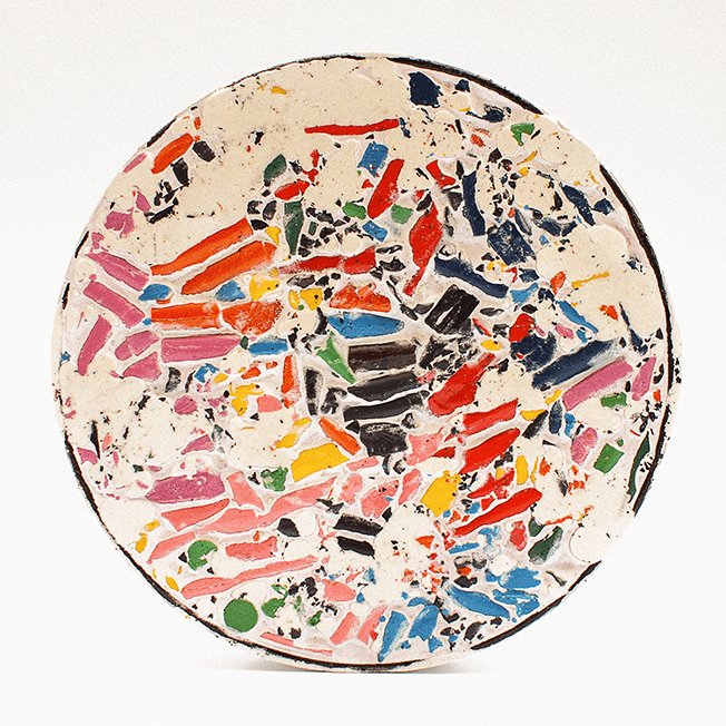 A round, mosaic-like disc made from crushed wax crayons encased in a light-colored filler, resembling a colorful, abstract pattern.