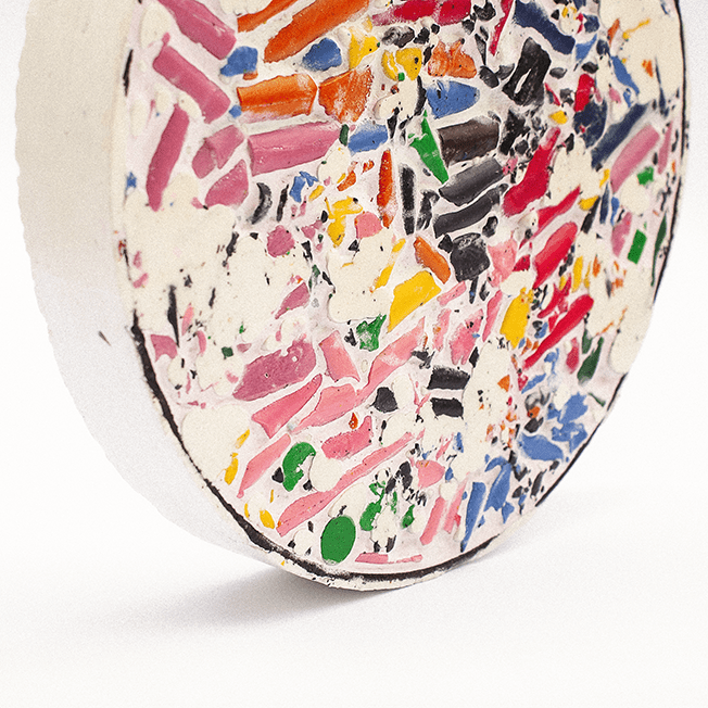 A colorful mosaic disc made from crushed wax crayons embedded in a white filler, viewed at an angle.