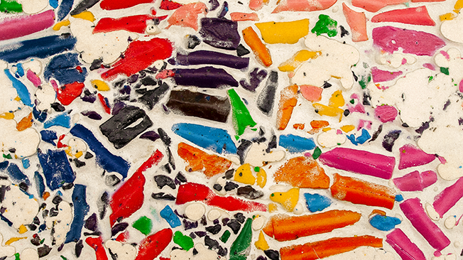 Close-up of a colorful mosaic made from crushed wax crayons set in a light filler, showcasing vibrant reds, blues, greens, and yellows.