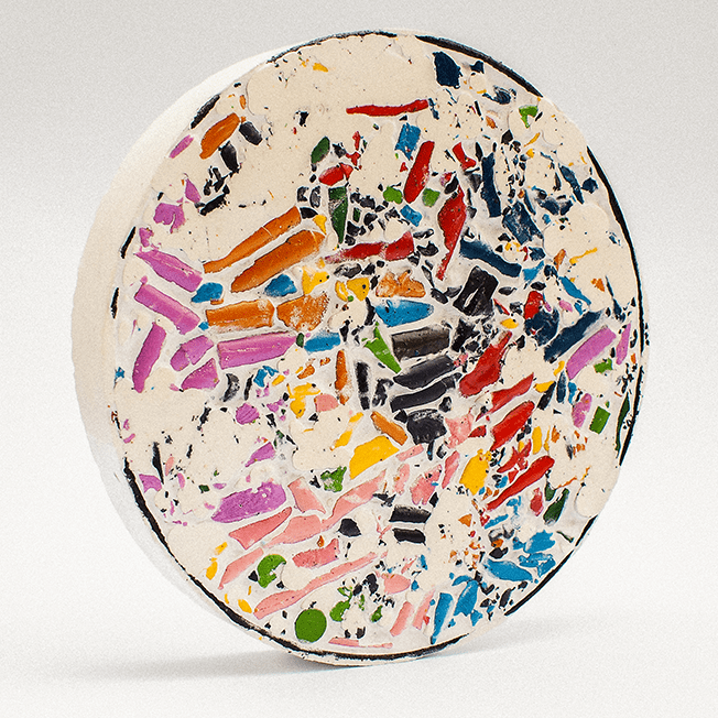 A colorful mosaic disc made from crushed wax crayons, set in a white resin-like filler, resembling the size of a dinner plate.