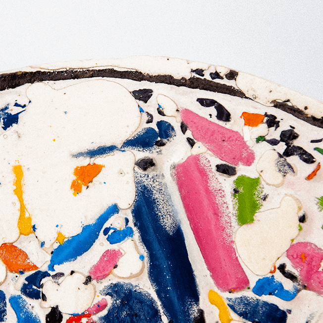 Close-up of a circular mosaic made from crushed wax crayons, set in a metal hoop, featuring vibrant colors like pink, blue, green, and orange.