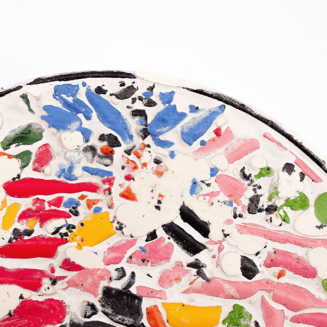 Close-up of a circular mosaic art piece made from crushed wax crayons set in a white filler, featuring various bright colors like blue, red, yellow, pink, and green.