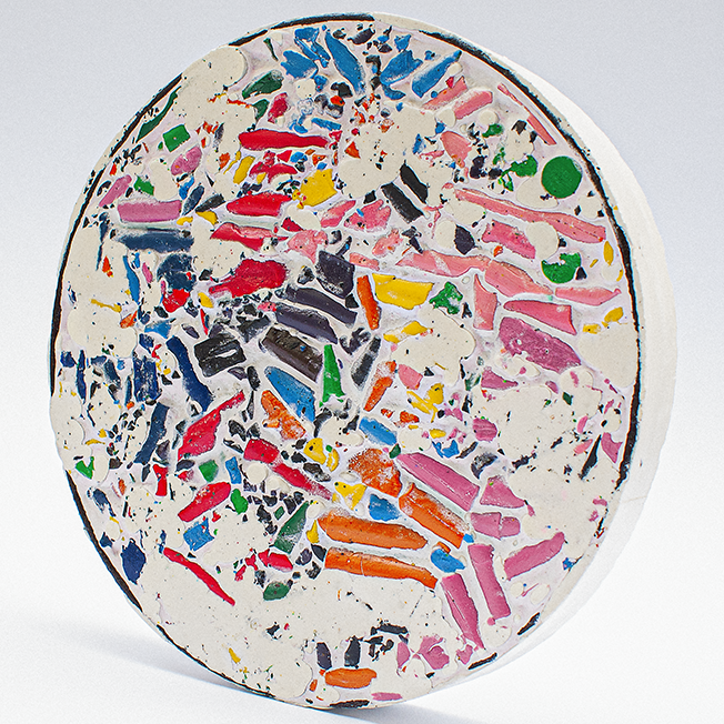 A round, colorful mosaic made from crushed wax crayons, set in a white plaster-like material, resembling the size of a dinner plate.