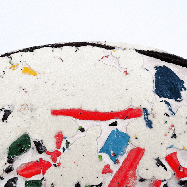 Close-up of a circular disc made from crushed crayons and filler, showing a mosaic-like texture with various colors including red, blue, green, yellow, and black, set against a white background.
