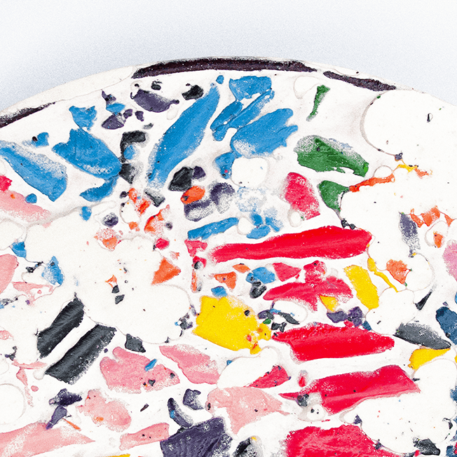 Close-up view of a colorful mosaic made from crushed wax crayons, arranged in a circular shape with a smooth white base.