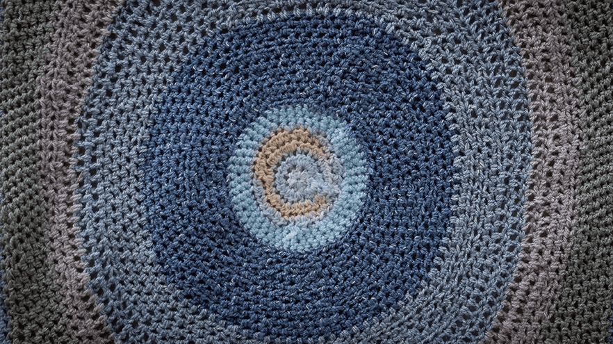 A close-up view of a crocheted piece featuring concentric circles in various shades of blue, grey, and a hint of beige, creating a soothing pattern.