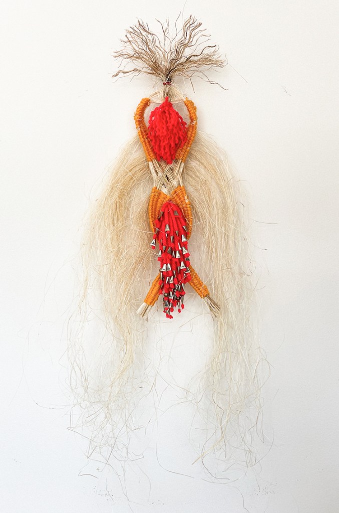 A woven textile artwork featuring vibrant red and orange elements, with long, flowing natural fibers creating an ethereal appearance.