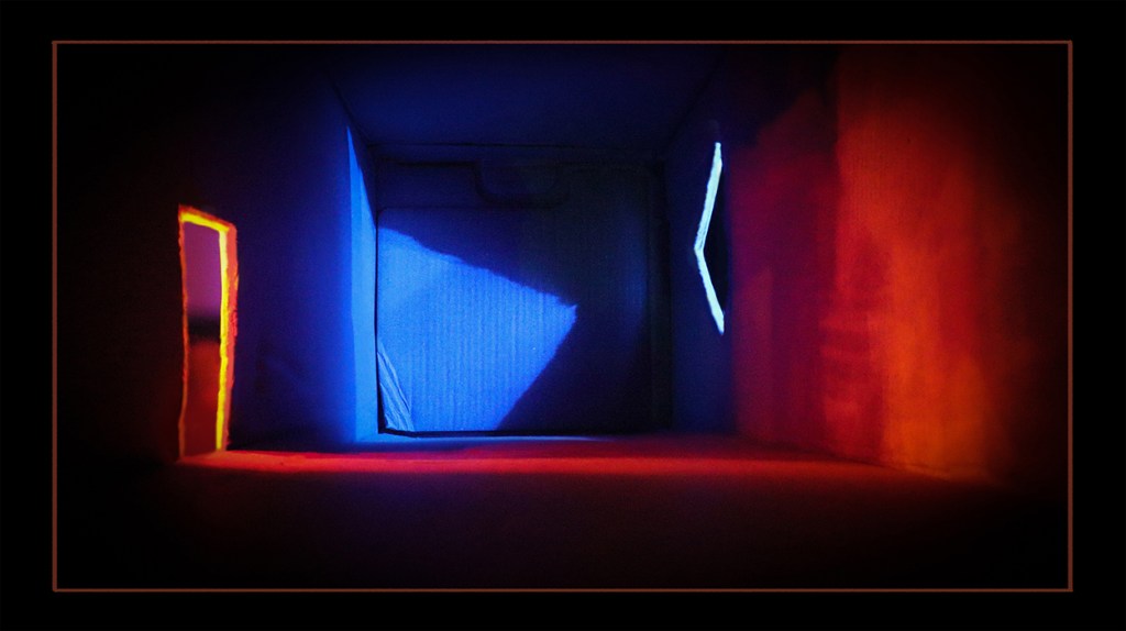 A cardboard model room illuminated with colorful lights, showcasing a doorway and geometric shapes on the walls.