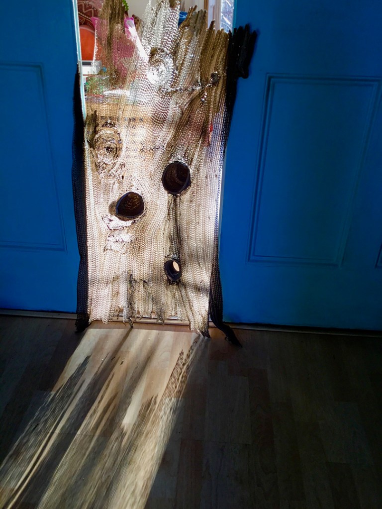An artistic depiction of light streaming through a cut-out piece resembling a tree log, positioned in a doorway with a blue door in the background. The shadows and textures created by the light highlight the natural patterns of the cut-out.