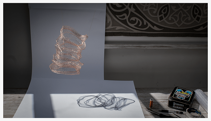 A copper mesh sculpture suspended over a white background, casting shadows on a surface covered with tools and a sketch. The setting features patterned curtains in the background.