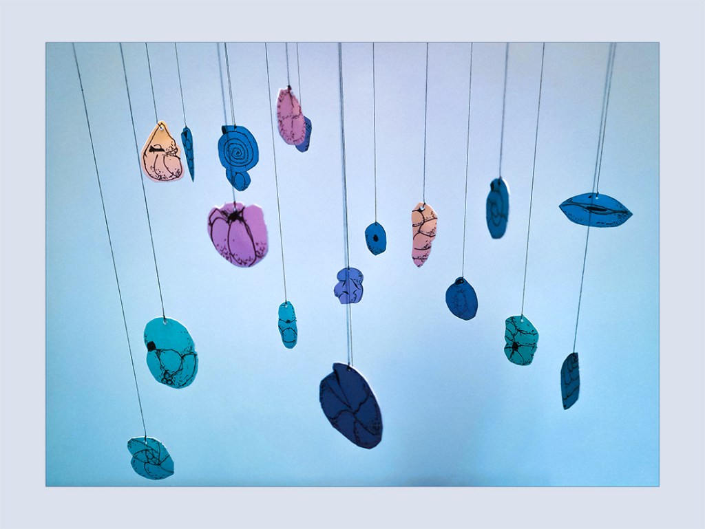 Colorful mobile featuring cut-out shapes inspired by foraminifera, suspended from thin threads against a softly lit background.