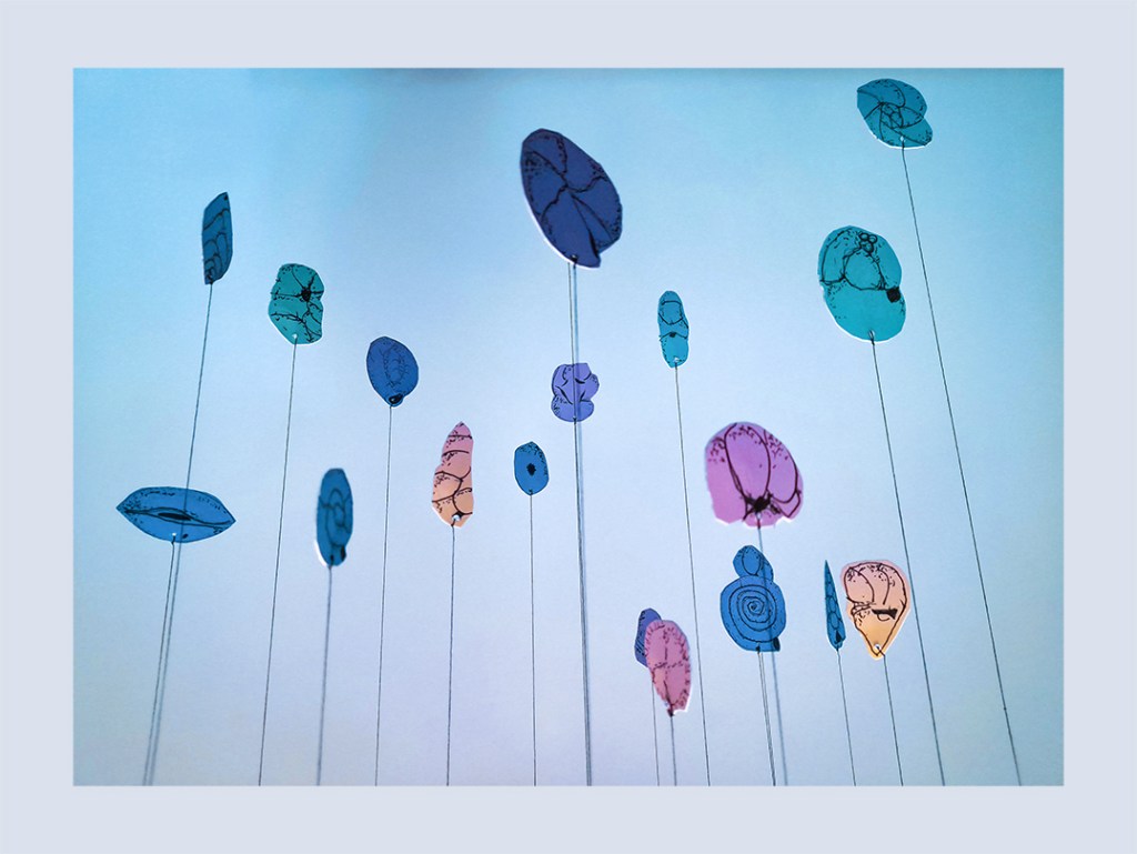 A display of colorful cut-outs representing foraminifera, suspended on thin strings against a light blue background.