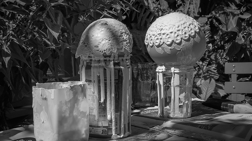 Two spherical shapes covered in white PVA glue and fabric doilies, resting on jars, with glue dripping down. The background features greenery, and the image is in black and white.