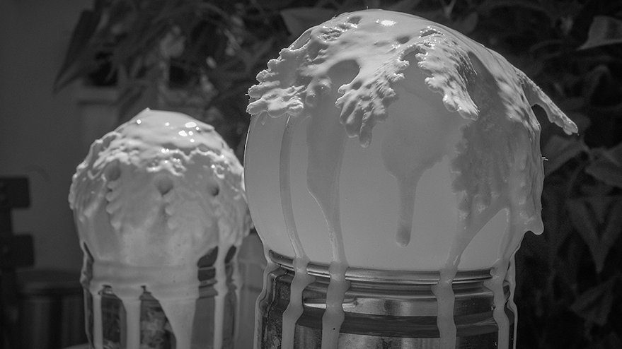Two spherical shapes covered with dripping white substance, resembling a frothy, textured coating, placed on metallic containers. In the background, leafy plants are visible.