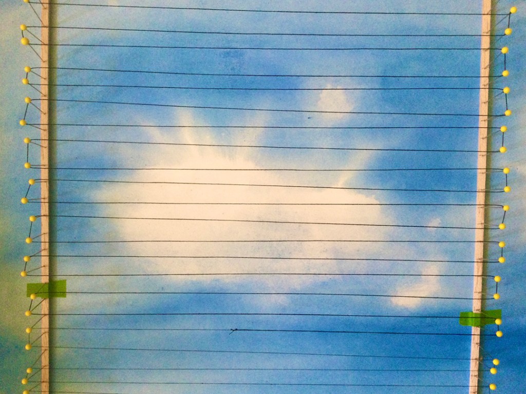 An abstract artwork depicting a sky with light clouds, featuring horizontal lines and yellow pins creating a visual structure, reminiscent of weaving patterns.