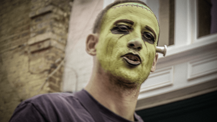 A person with green face paint and dark makeup, resembling a monster, stands in front of a brick wall, likely during a carnival event.