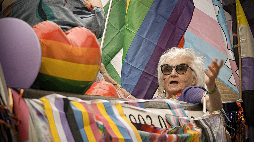 An elderly woman with glasses joyfully participates in a parade, surrounded by colorful flags and decorations, including rainbow hearts and striped banners.