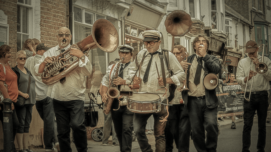 A brass band playing in a parade during the carnival in Whitstable, featuring musicians in formal attire and a crowd of spectators behind them.