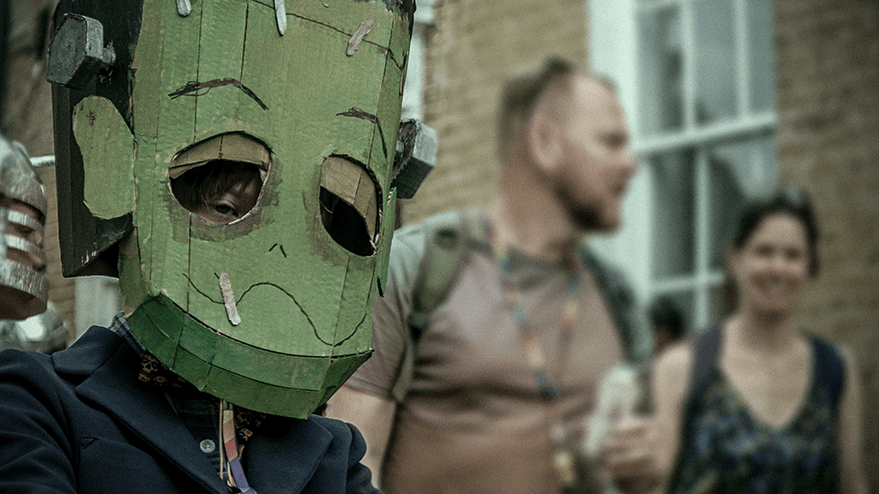 A child wearing a large cardboard Frankenstein mask, surrounded by adults in the background during a carnival celebration.