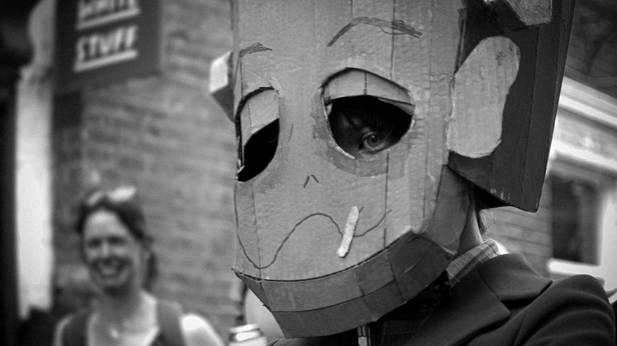 A person wearing a cardboard mask with cartoonish facial features, standing in a carnival setting. In the background, another person is smiling.