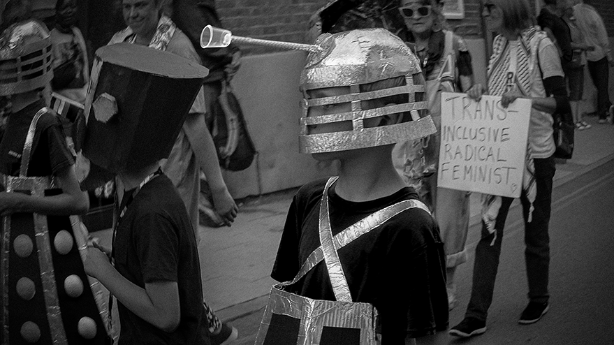 Children participating in a carnival parade wearing cardboard Dalek costumes, with one holding a sign that reads 'TRANS INCLUSIVE RADICAL FEMINIST'.