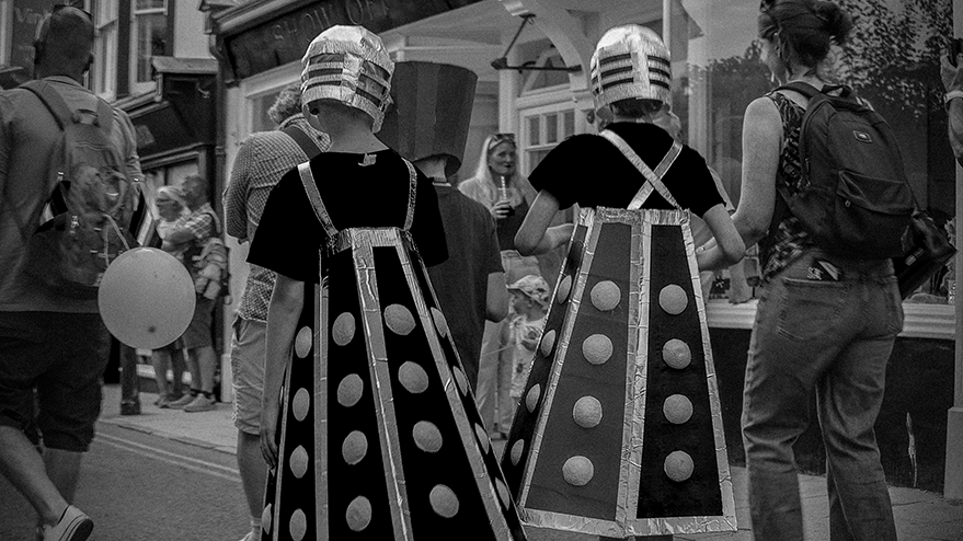Two children wearing cardboard Dalek costumes with silver helmets and polka-dotted capes, walking through a crowd during a carnival parade.