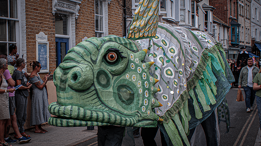 A large, colorful fish puppet being paraded on a street during the Whitstable carnival, with onlookers watching the event.