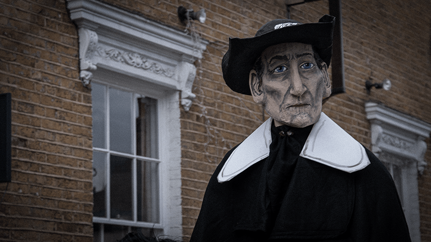 A puppet figure dressed in black with a large hat, resembling a historical character, stands against a brick wall with windows in the background.