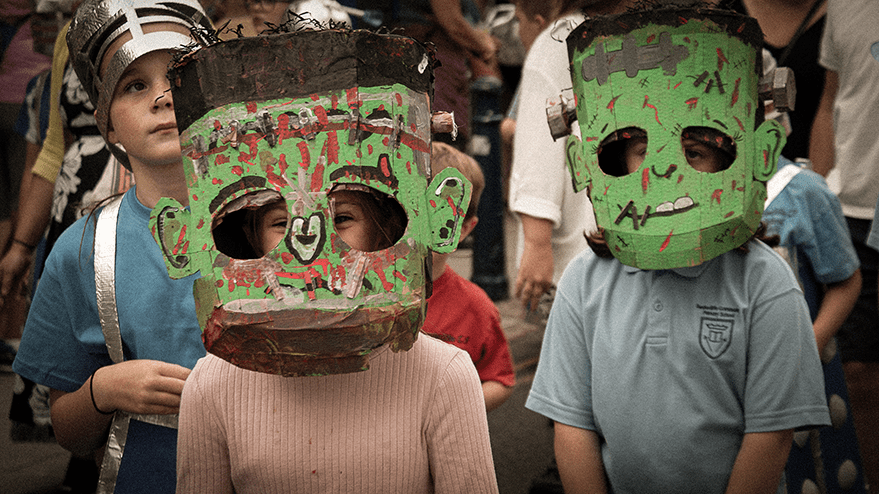 Two children wearing colorful cardboard monster masks, with one mask resembling Frankenstein, during a carnival parade.