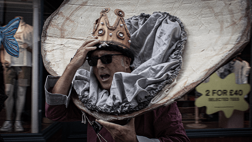 A man wearing sunglasses and a crown, holding a large, decorative collar while expressing excitement during the Whitstable carnival.