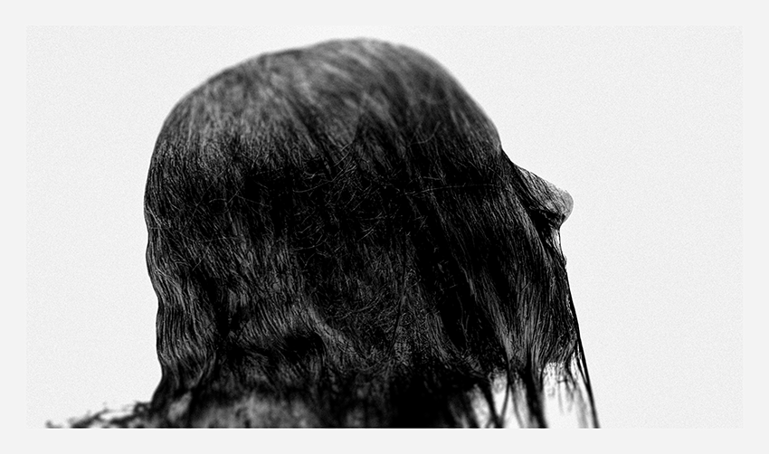 A black and white image of a person's head facing sideways, with long, dark hair partially covering the face.