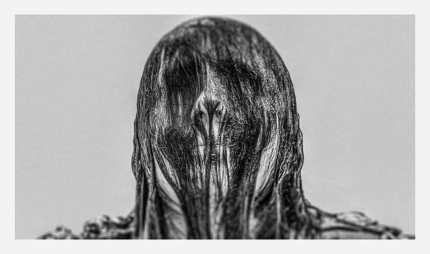 A close-up, high-contrast black and white portrait of a figure with long, flowing hair, partially obscuring the face, creating a haunting and mysterious appearance.