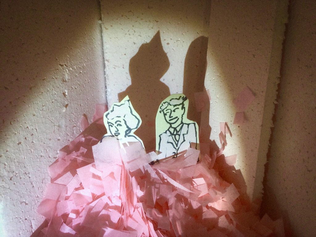 Two cartoon-like paper cutouts of a man and a woman standing on a pile of pink shredded paper, with shadows cast on a textured wall.